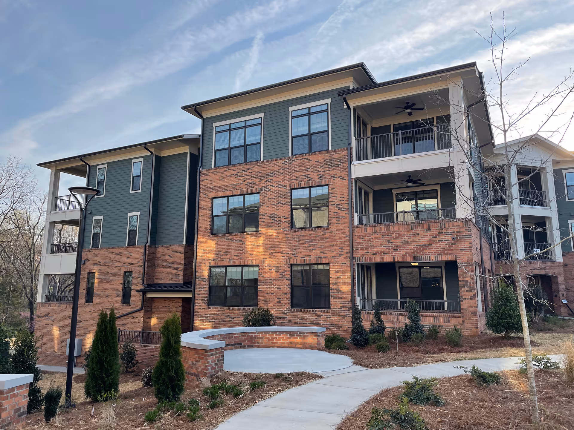 Three-story brick and green-siding apartment building with covered balconies and a curved paved walkway in front.
