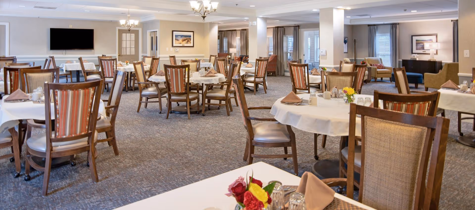A spacious dining room with multiple round tables covered with white tablecloths, each set with napkins, glasses, and small floral centerpieces. The room features wooden chairs with striped cushions, carpeted flooring, and soft lighting from chandeliers. In the background, there is a large flat-screen TV mounted on the wall and a seating area with armchairs and a sideboard with a lamp.