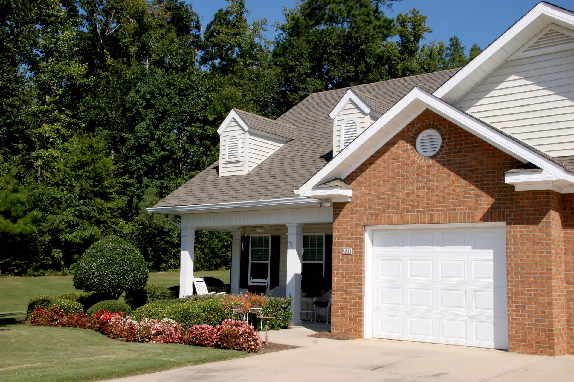 Exterior view of a single-story brick and siding building with a white garage door, a covered porch with white columns, manicured bushes, and colorful flowers in front. Tall green trees are visible in the background under a clear blue sky.