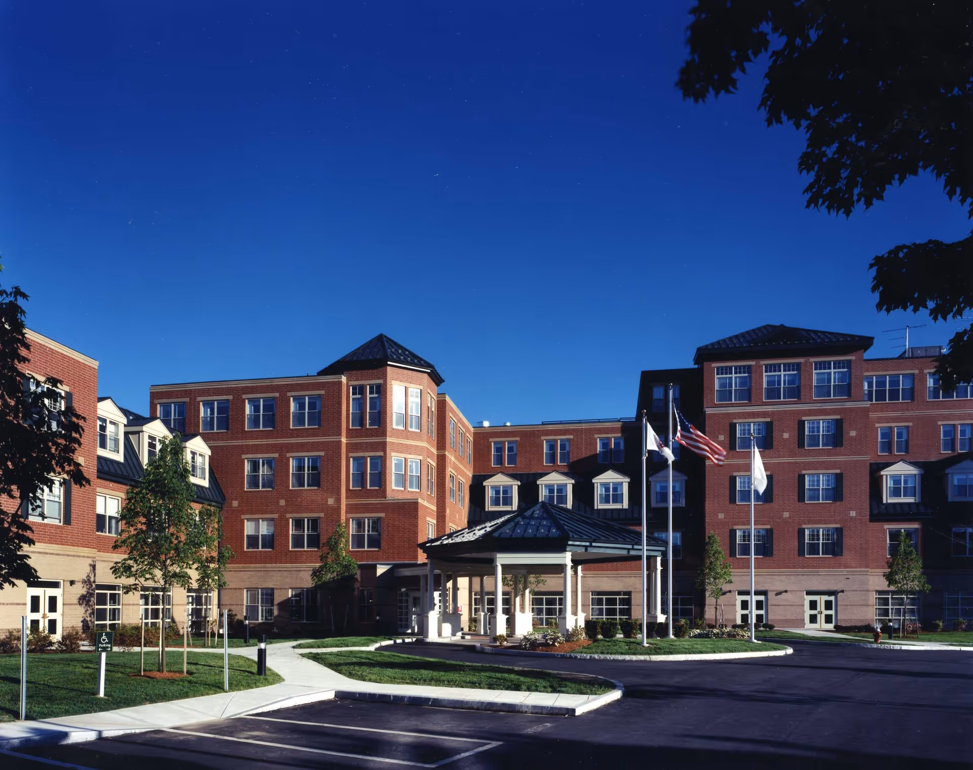Exterior view of a multi-story brick building with a covered entrance and three flagpoles displaying flags, surrounded by a paved driveway, sidewalks, and small trees under a clear blue sky.