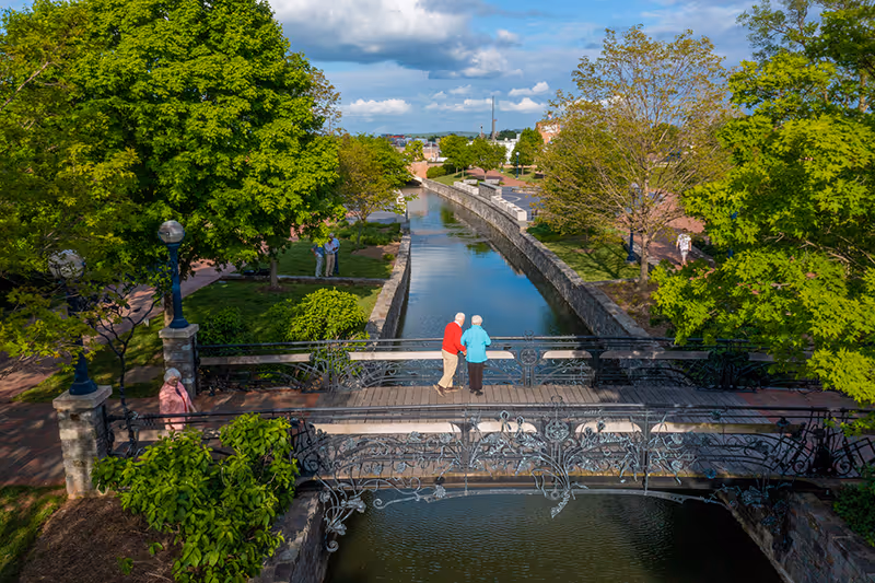 A scenic outdoor view of a narrow canal with a decorative iron bridge crossing over it. Two elderly people stand on the bridge looking at the water. The area is surrounded by green trees and walking paths with a few other people strolling nearby under a partly cloudy sky.