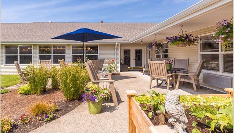 Sunny outdoor patio courtyard with wooden chairs and tables, potted plants, hanging baskets, and a blue umbrella in front of a single-story building.