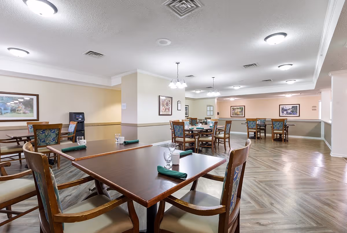 A spacious dining room in a senior living facility with several wooden tables and chairs arranged neatly. Each table is set with glasses, cups, and green napkins. The room has light-colored walls adorned with framed artwork, wood-patterned flooring, and ceiling lights providing bright illumination.