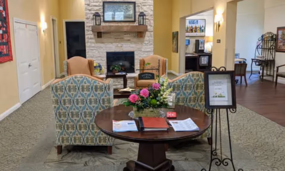 A senior living facility lounge with patterned sofas and chairs arranged around a round table with a floral centerpiece in front of a stone fireplace.