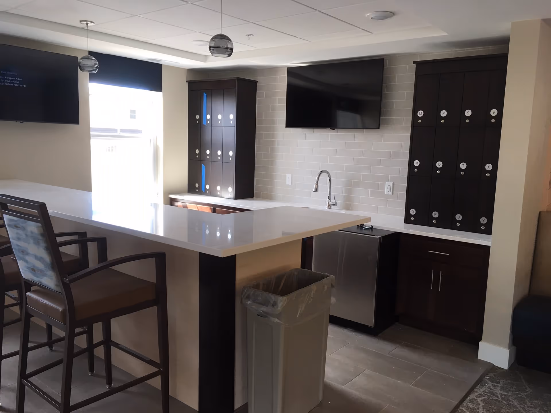 Interior view of a modern kitchen area in a senior living facility with a white countertop island, two high chairs, a stainless steel sink, dark wood cabinets with labeled compartments, a mounted flat-screen TV, and a trash bin. There is a window with a black shade letting in natural light.