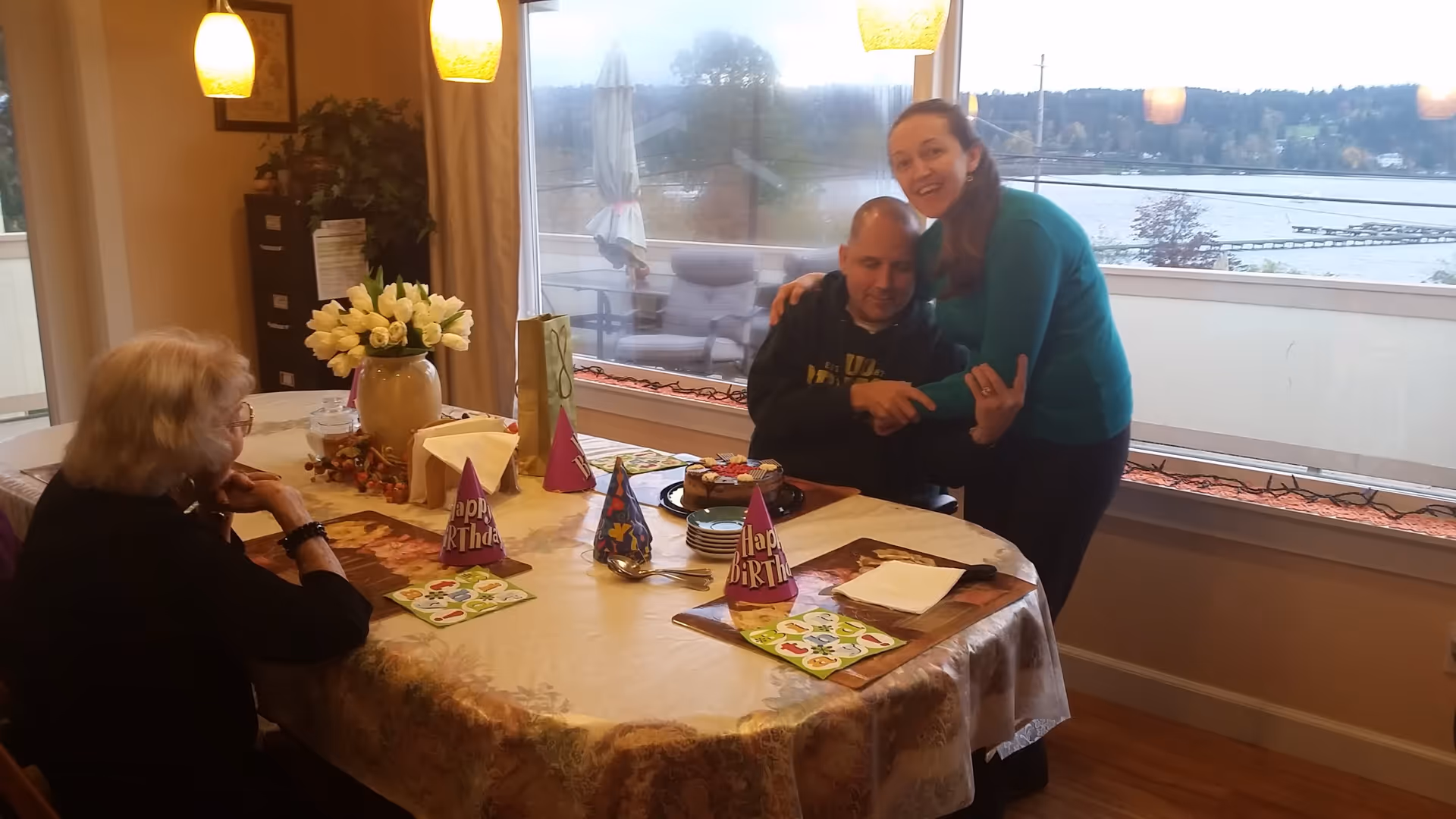 Three people gathered around a dining table decorated for a birthday celebration with party hats, a cake, and a vase of white tulips. The room has large windows showing a lake and outdoor seating area in the background.