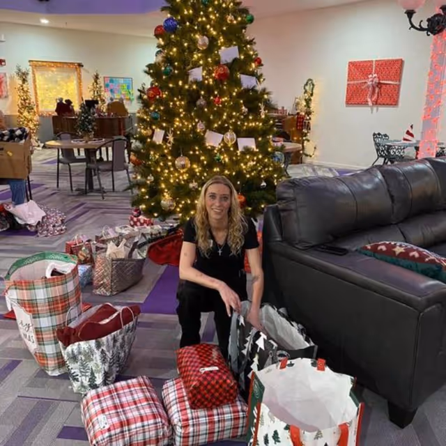 A woman with long blonde hair is kneeling on the floor in front of a decorated Christmas tree with lights and ornaments. She is surrounded by wrapped presents and gift bags. The setting appears to be a cozy indoor common area with a dark leather couch, tables, chairs, and holiday decorations on the walls.