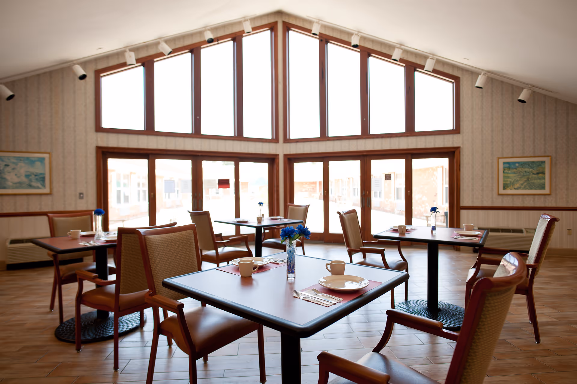 A bright dining room with several tables and chairs set for meals beneath large triangular windows and a vaulted ceiling.