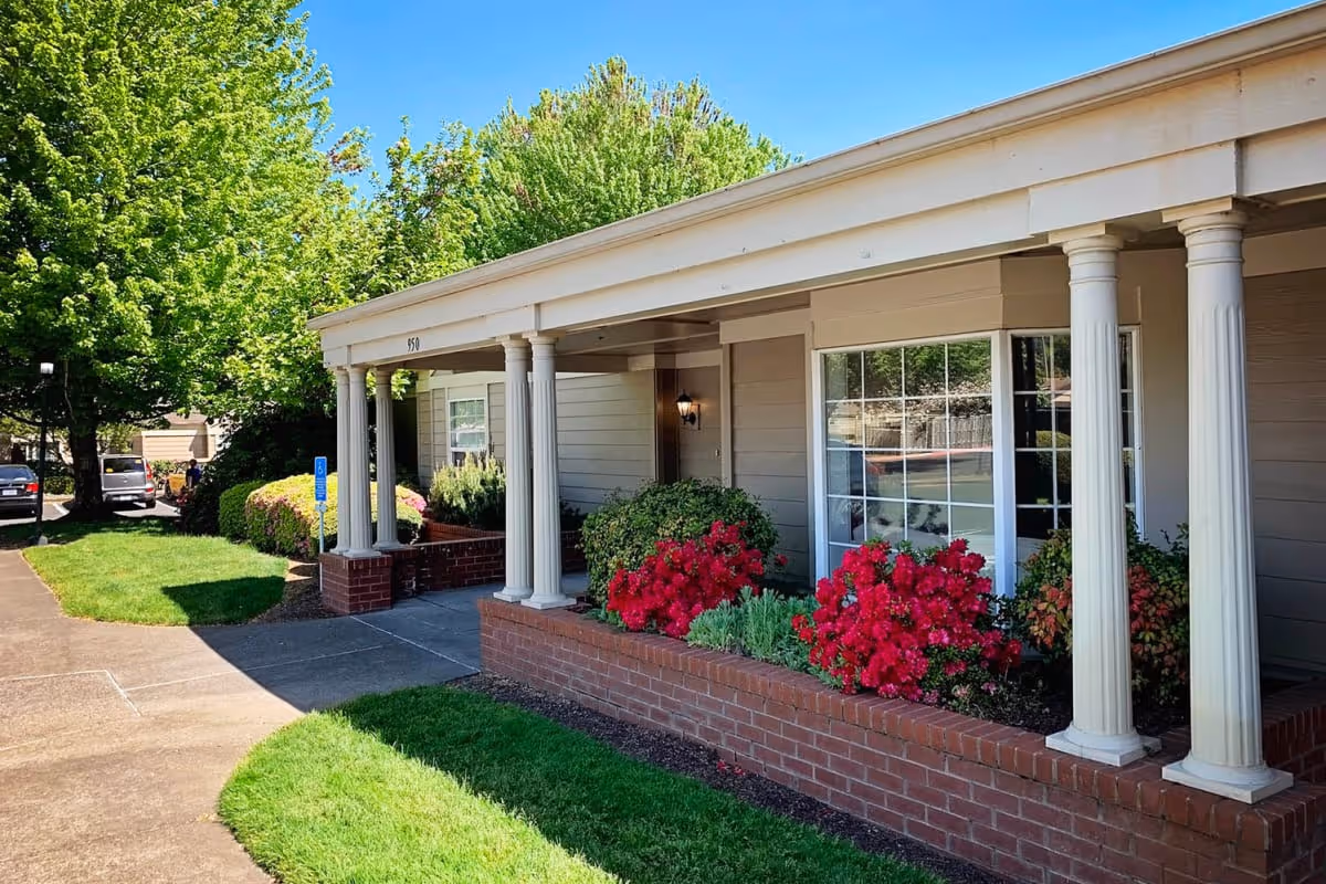Front entrance of a single-story senior living building with white columns, a brick planter and red flowering shrubs along a sidewalk.