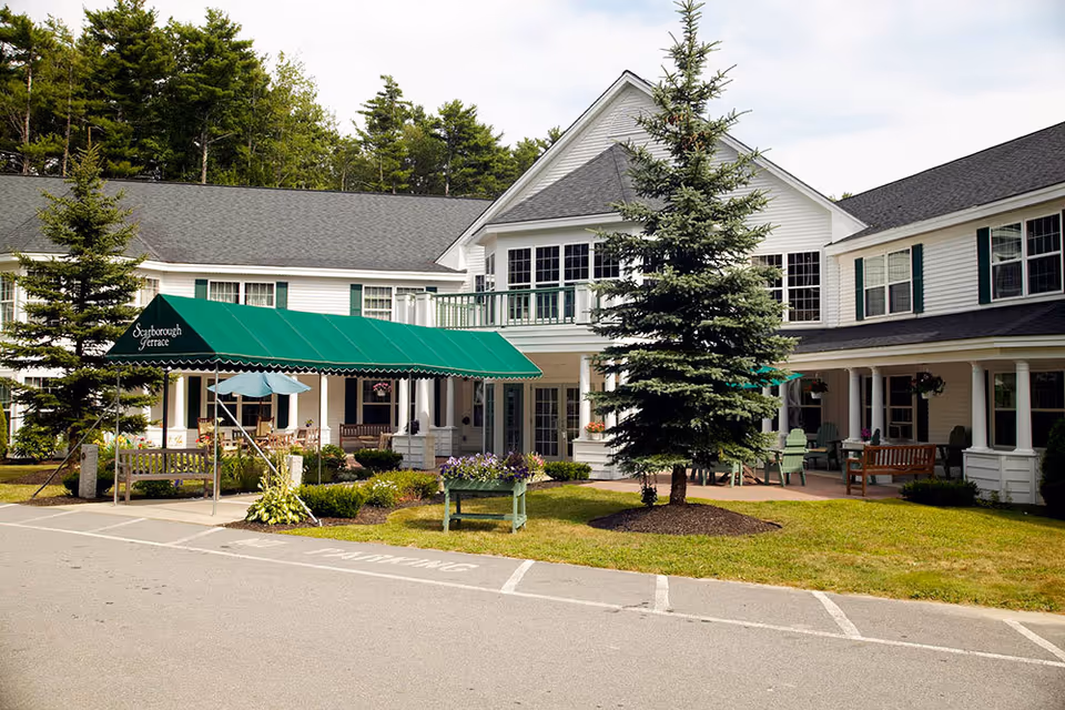 Front exterior of a white two-story senior living building with a green awning reading 'Scarborough Terrace', landscaped lawn, benches and patio seating.