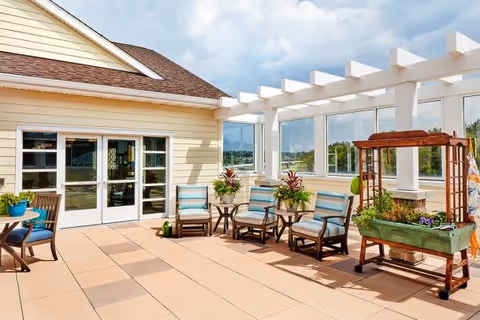Outdoor patio area at Alto Overland Park with cushioned chairs arranged around small tables, potted plants, a wooden planter on wheels with flowers, and a pergola structure with large windows overlooking greenery under a partly cloudy sky.