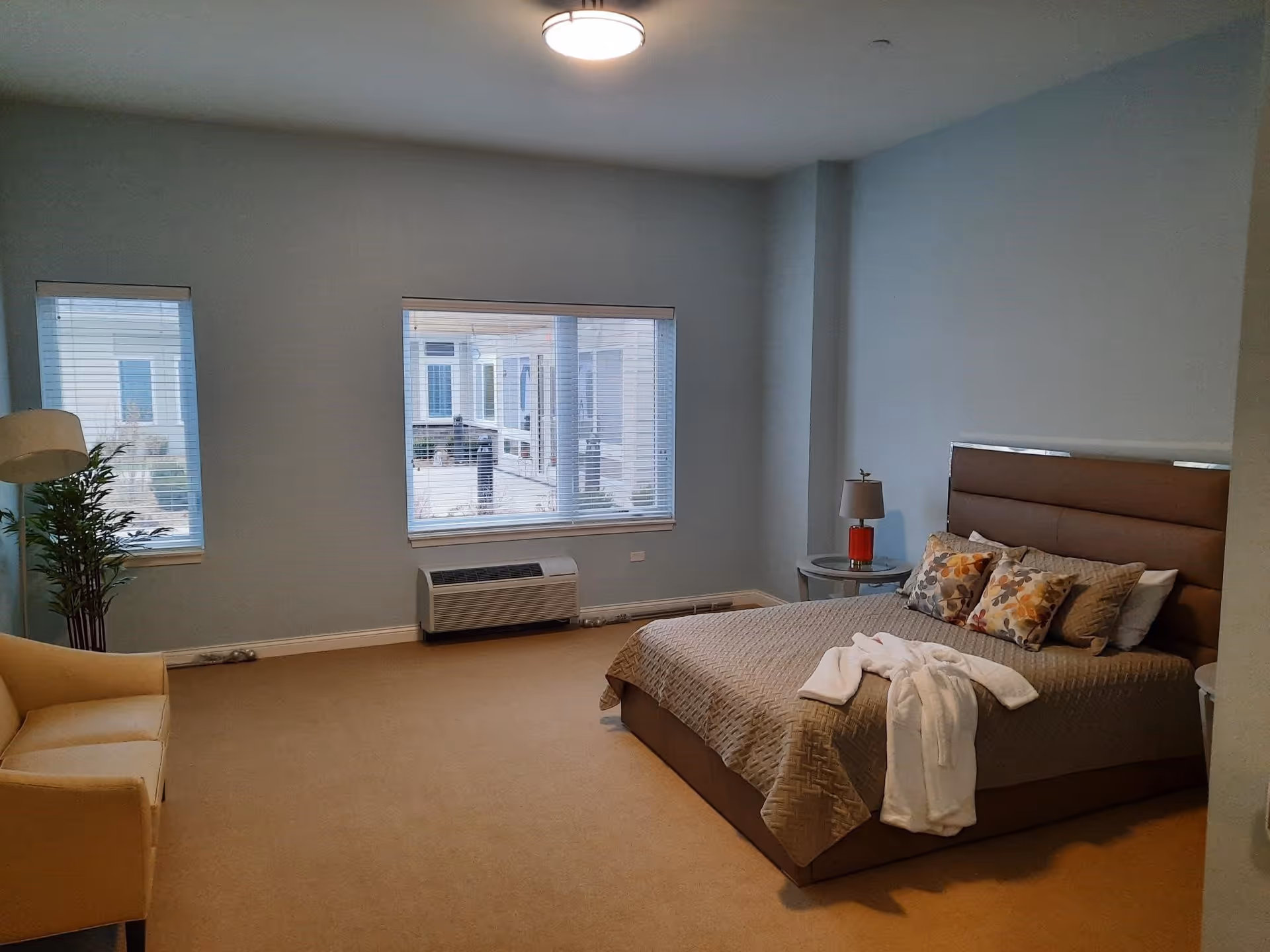 A bedroom with a large bed covered in a beige quilt and decorative pillows. A white robe is draped on the bed. There are two windows with blinds, a beige armchair, a floor lamp, a small round side table with a lamp, and a potted plant in the corner. The walls are painted light blue and the carpet is beige.