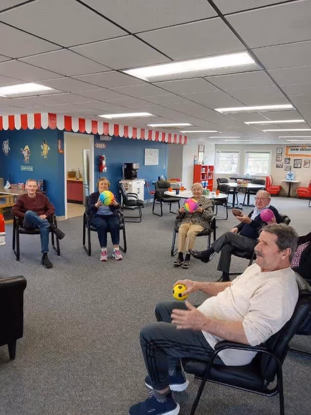 A group of elderly people sitting in a circle in a spacious, well-lit room with gray carpet and white ceiling tiles. They are holding colorful balls, appearing to participate in a seated group activity. The room has blue walls with a red and white striped canopy above a doorway, and various chairs and tables are visible in the background.