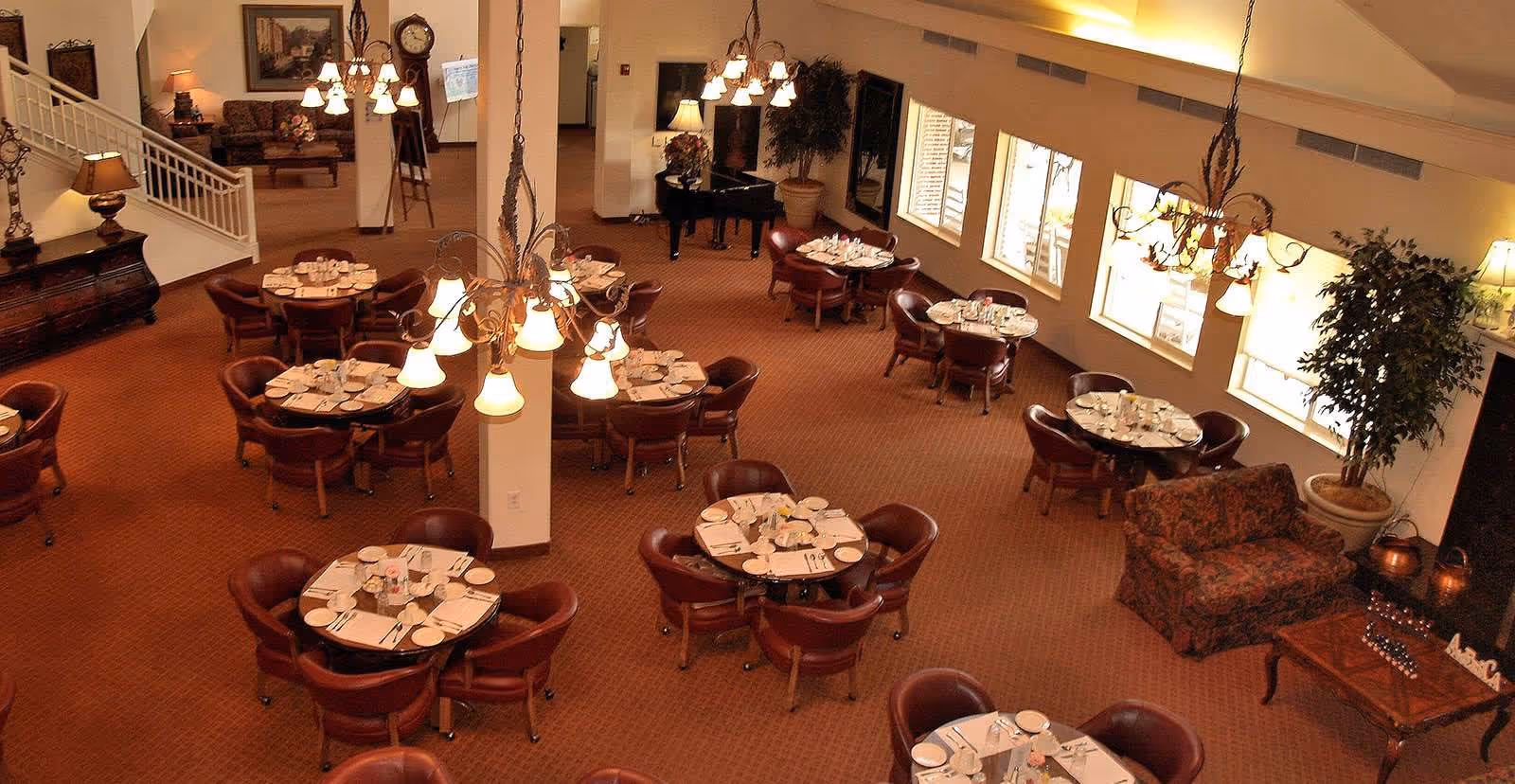 A spacious dining room with multiple round tables set with plates, glasses, and silverware. The room has warm lighting from chandeliers and table lamps, carpeted floors, and large windows letting in natural light. There is a piano in the corner and comfortable seating areas with armchairs and a small sofa. The decor includes plants and framed artwork on the walls.