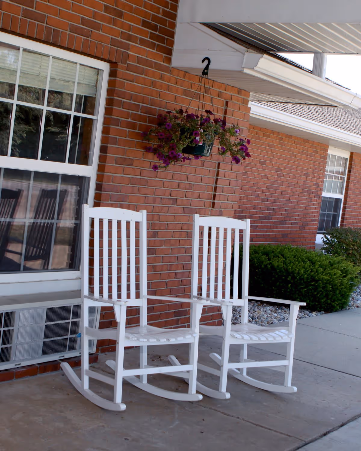 Two white wooden rocking chairs on a concrete porch in front of a red brick wall with a window and an air conditioning unit below it. A hanging basket with purple flowers is suspended above the chairs. Green bushes and a sidewalk are visible to the right.