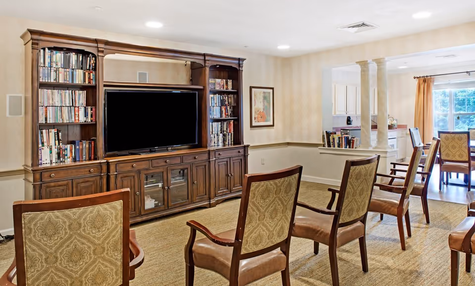 A cozy living room area with a large wooden entertainment center filled with books and a flat-screen TV. Several upholstered chairs with wooden frames are arranged facing the TV. The room has beige walls, carpeted floor, and recessed ceiling lights. In the background, there is a dining area with a table and chairs near a window with curtains.