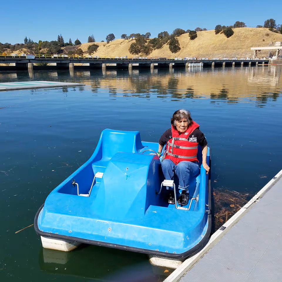 An elderly woman wearing a red life jacket is sitting on a blue pedal boat on a calm body of water near a dock. In the background, there is a bridge and dry, grassy hills under a clear blue sky.