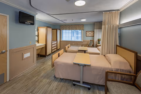 A senior living facility bedroom with two hospital-style beds, each equipped with an overbed table. The room has light blue walls, wood paneling, a wall-mounted TV, a window with blinds and a valance, a wooden dresser, a sink with a mirror, and a privacy curtain between the beds. There is also a chair next to one of the beds.