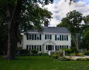 A two-story white house with green shutters surrounded by trees and greenery under a partly cloudy sky.