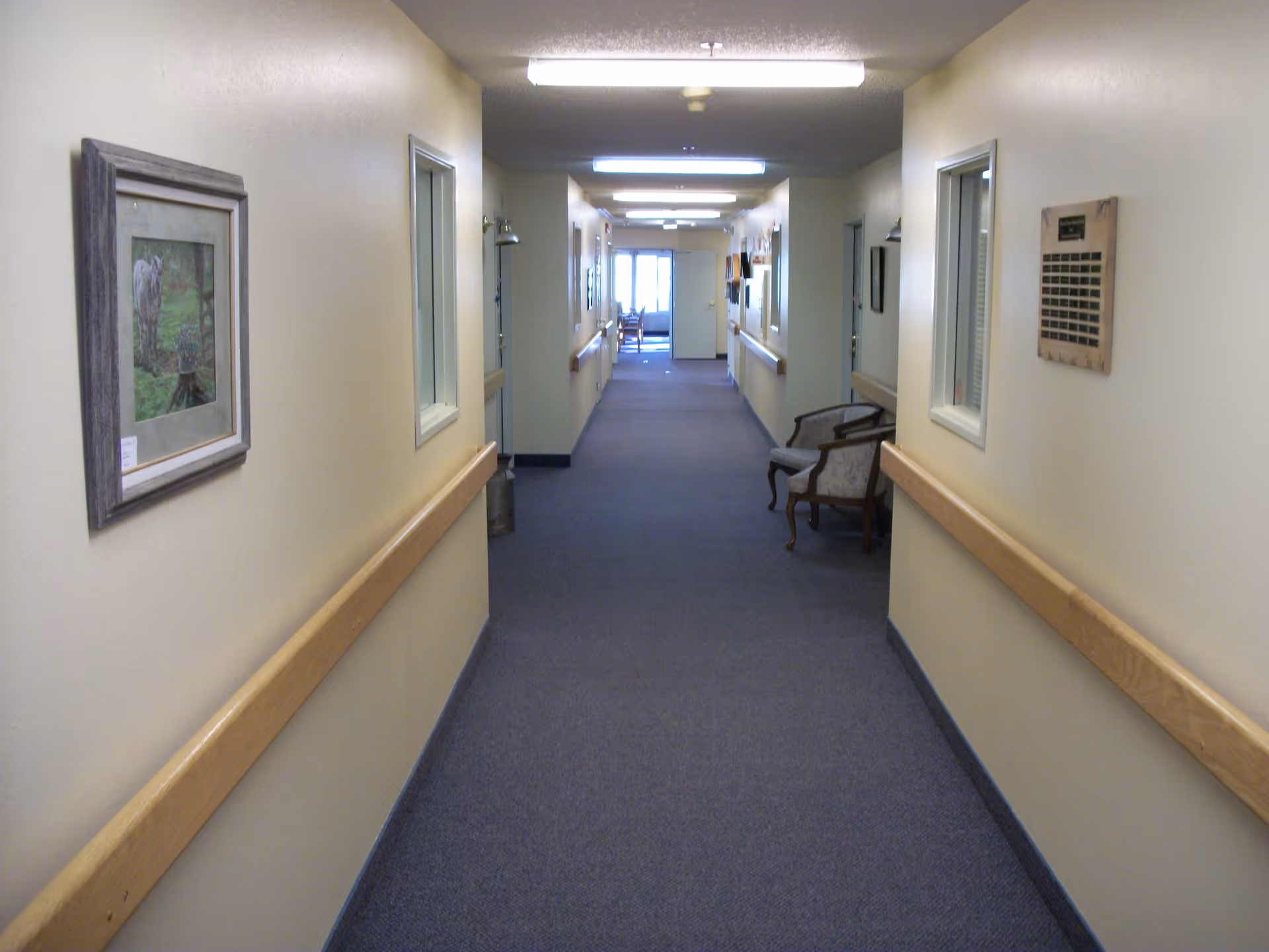 Carpeted interior hallway of a retirement center with handrails, framed artwork and chairs along the walls leading to a distant window.