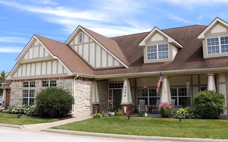 Exterior view of a single-story senior living facility building with stone and beige siding, a brown shingled roof, and a covered porch with white columns. The porch has chairs, potted plants, and an American flag. The lawn is well-maintained with bushes and flowers.