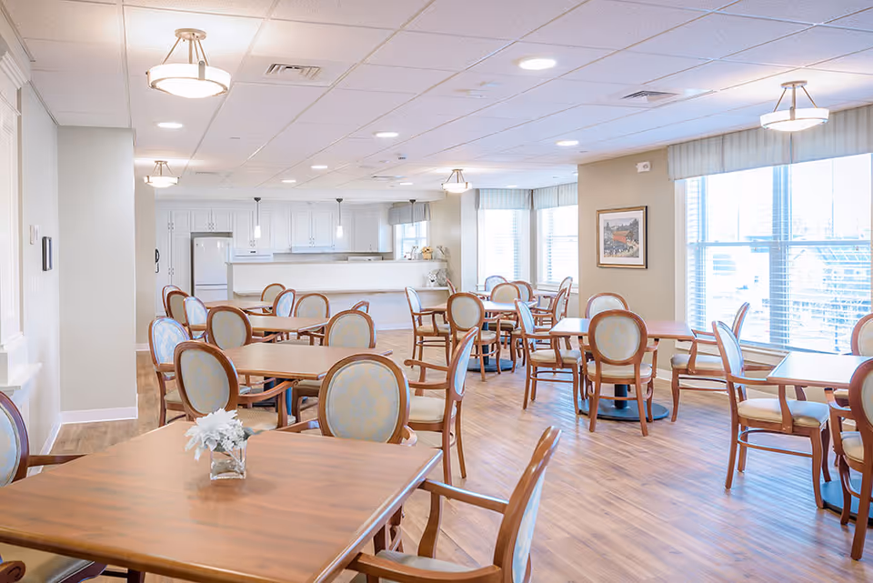 A bright and spacious dining room with multiple wooden tables and upholstered chairs arranged neatly. Large windows with blinds allow natural light to fill the room. The back of the room features a kitchen area with white cabinetry and pendant lights hanging over the counter.