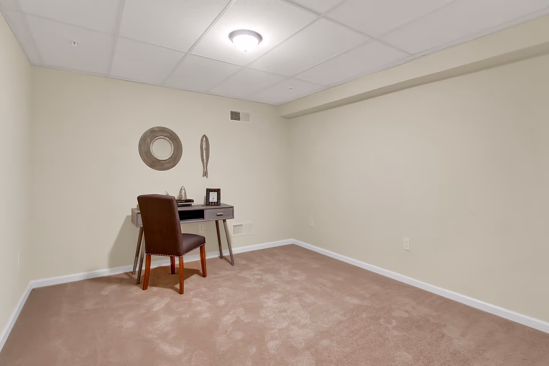 A small room with beige carpet and cream-colored walls, featuring a simple desk with a brown chair. On the wall above the desk are two decorative items: a round mirror and a fish-shaped wall hanging. The ceiling has white tiles with a single light fixture.