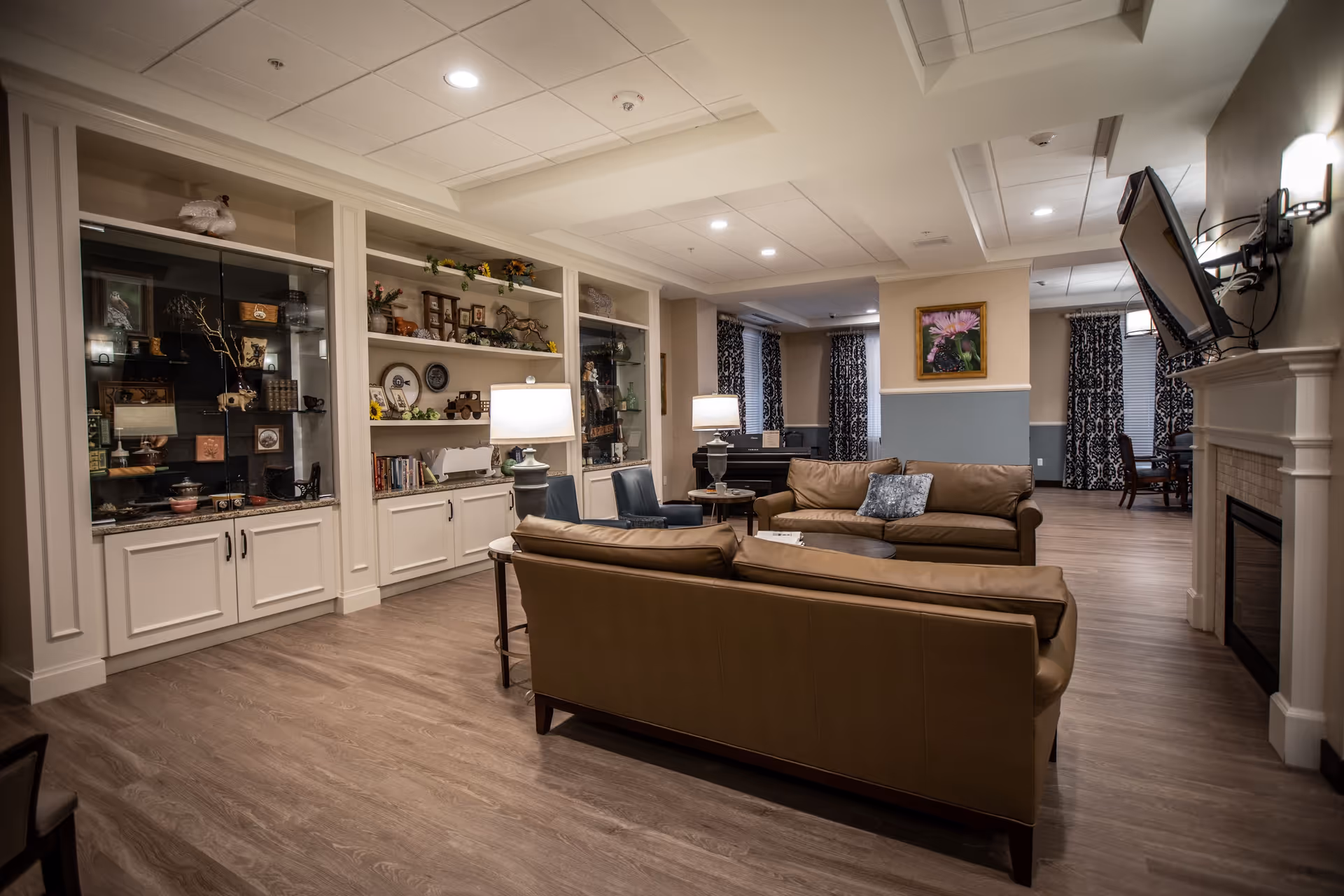 A cozy living room area in a senior living facility with two brown leather sofas, two blue chairs, a round coffee table, and two table lamps. The room features built-in shelves and cabinets filled with decorative items, a wall-mounted TV above a fireplace, and patterned curtains on the windows. The flooring is wood-style, and the walls are painted in neutral tones with a blue accent.
