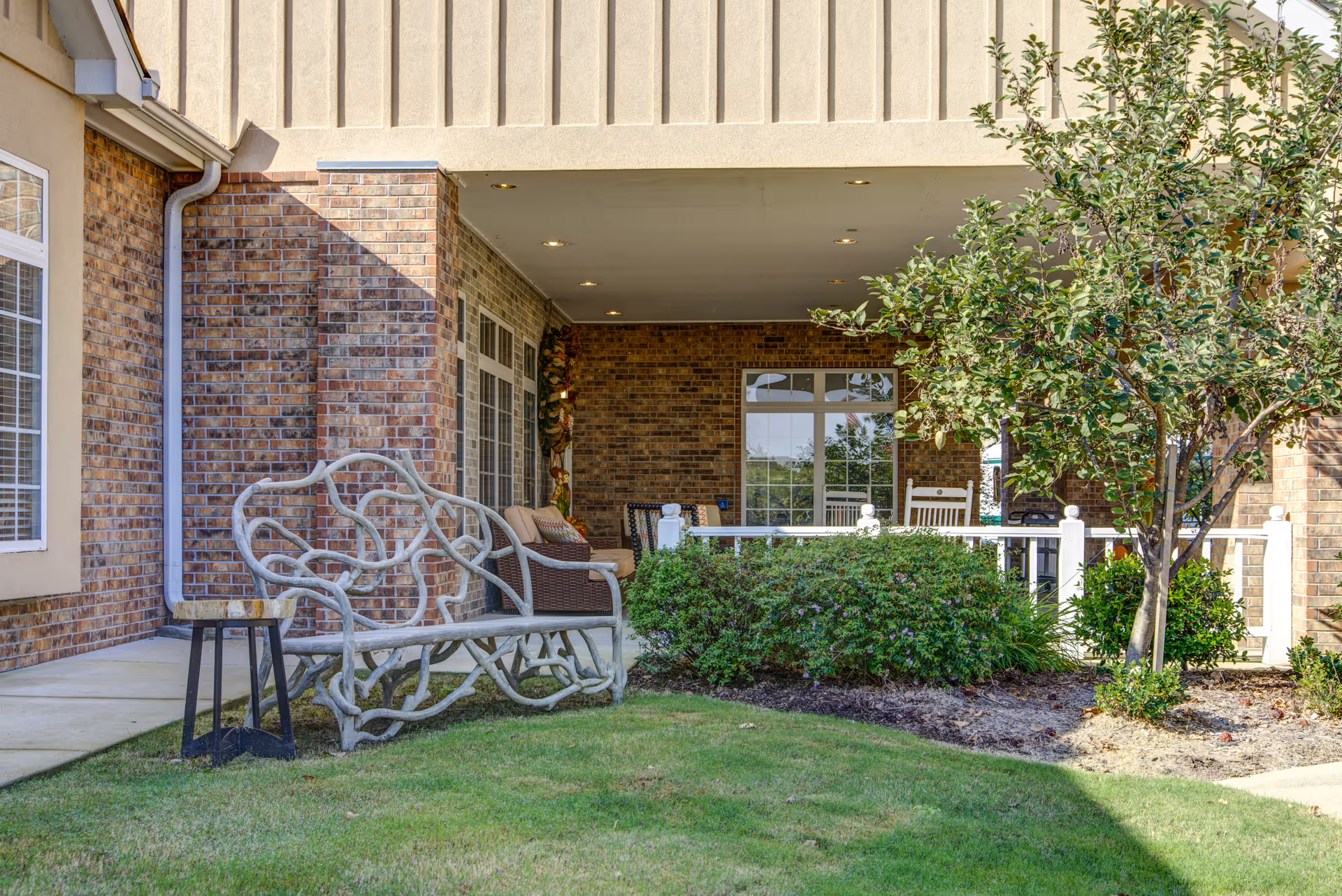 Outdoor patio area of a senior living facility with a decorative metal bench, a small side table, green bushes, a tree, and a covered porch with wicker furniture and rocking chairs against a brick wall.