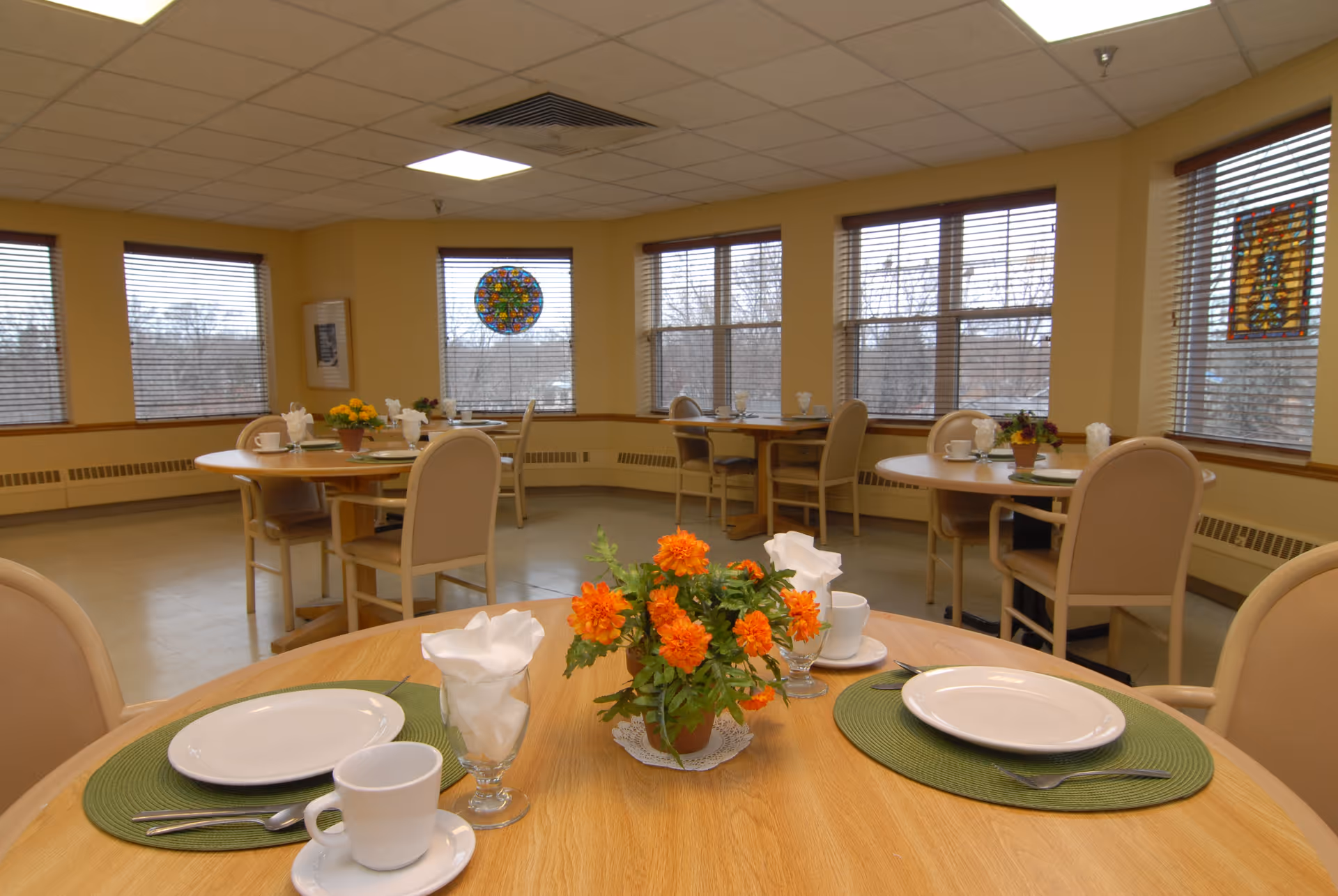 A dining room in a senior living facility with round wooden tables set with white plates, cups, silverware, and napkins. Each table has a small flower arrangement with orange flowers. The room has large windows with blinds and stained glass decorations, beige walls, and a tiled floor.