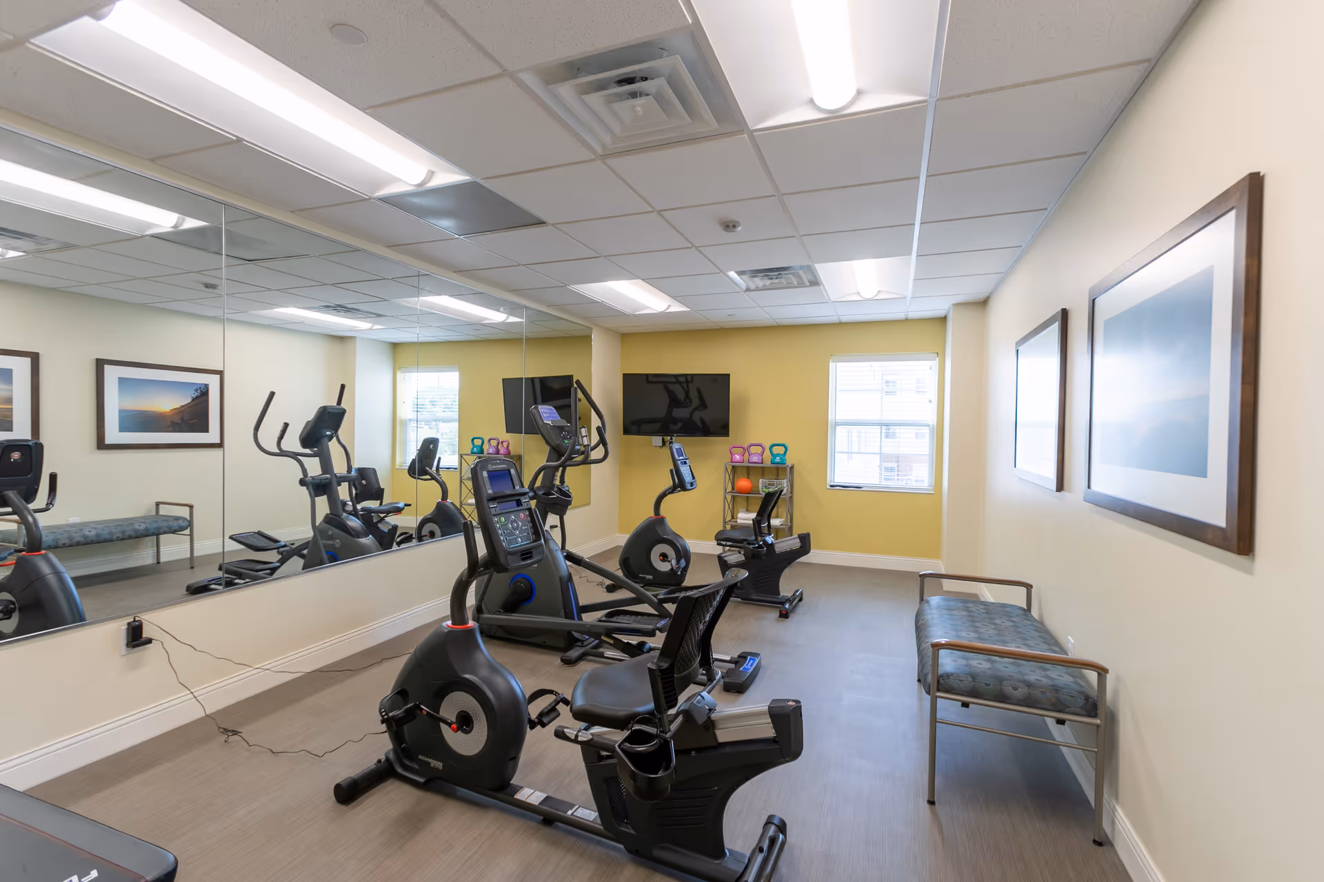 A small fitness room with exercise bikes, an elliptical machine, a bench, and a rack with colorful kettlebells and an exercise ball. The room has a large mirror on one wall, two windows, framed pictures on the walls, and a wall-mounted TV.