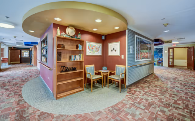 A cozy corner in a senior living facility hallway featuring a small wooden bookshelf with books and decorative items, a wooden table with two chairs, and framed artwork on the walls. The hallway has patterned carpet and leads to other rooms with open doors.