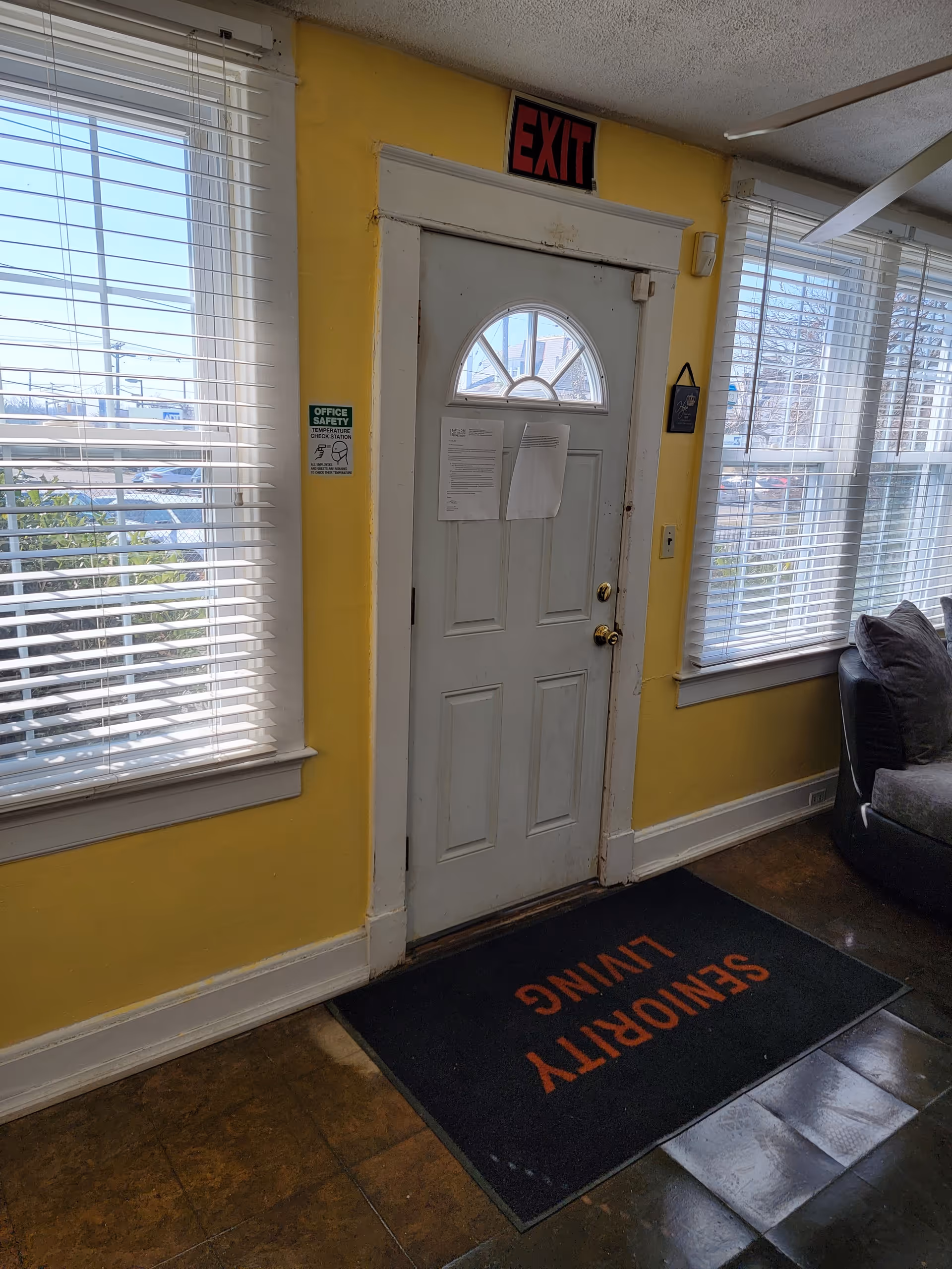 Interior view of a room with yellow walls featuring a white exit door with a semicircular window at the top. There are two large windows with white blinds on either side of the door. A black mat with the words 'SENIORITY LIVING' in orange is placed in front of the door. A gray couch with pillows is partially visible on the right side.