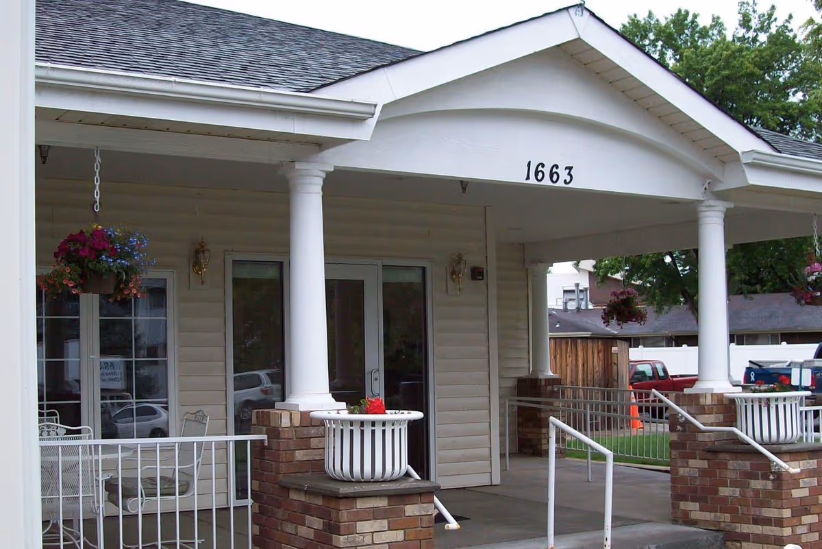 Entrance of a building with the number 1663 displayed above the doorway, featuring white columns, brick pillars with white planters containing flowers, hanging flower baskets, and a small patio area with white metal chairs and a table.