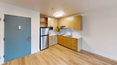 Studio kitchenette with light wood cabinets, stainless steel refrigerator and sink, and a blue entry door.