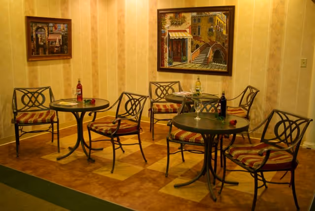 A small seating area with three round tables and six metal chairs with striped cushions. Each table has a bottle of wine and a single rose placed on it. The walls are decorated with two framed paintings depicting street scenes.