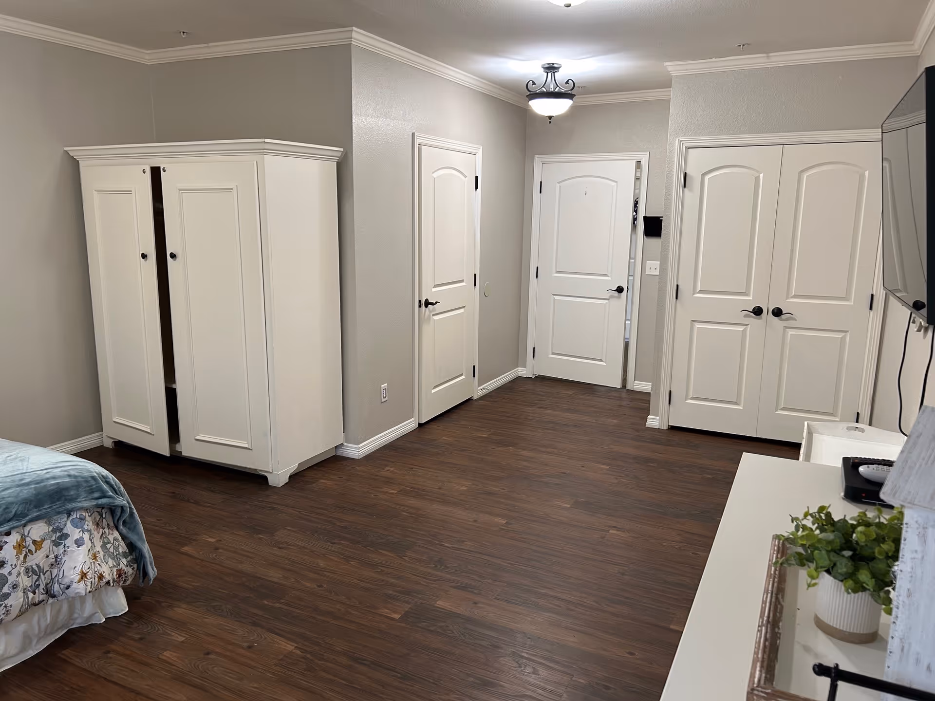 Spacious bedroom with wood floors, a white wardrobe and dresser, multiple doors, a partially visible bed, and a wall-mounted TV.