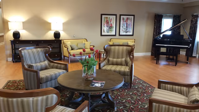 A cozy living room area with four striped armchairs arranged around a round wooden coffee table with a vase of red tulips and a magazine. In the background, there are two yellow sofas, a black grand piano, two framed floral artworks on the wall, two table lamps on a dark wooden console, and windows with dark curtains. The floor is wooden with a patterned area rug under the seating area.