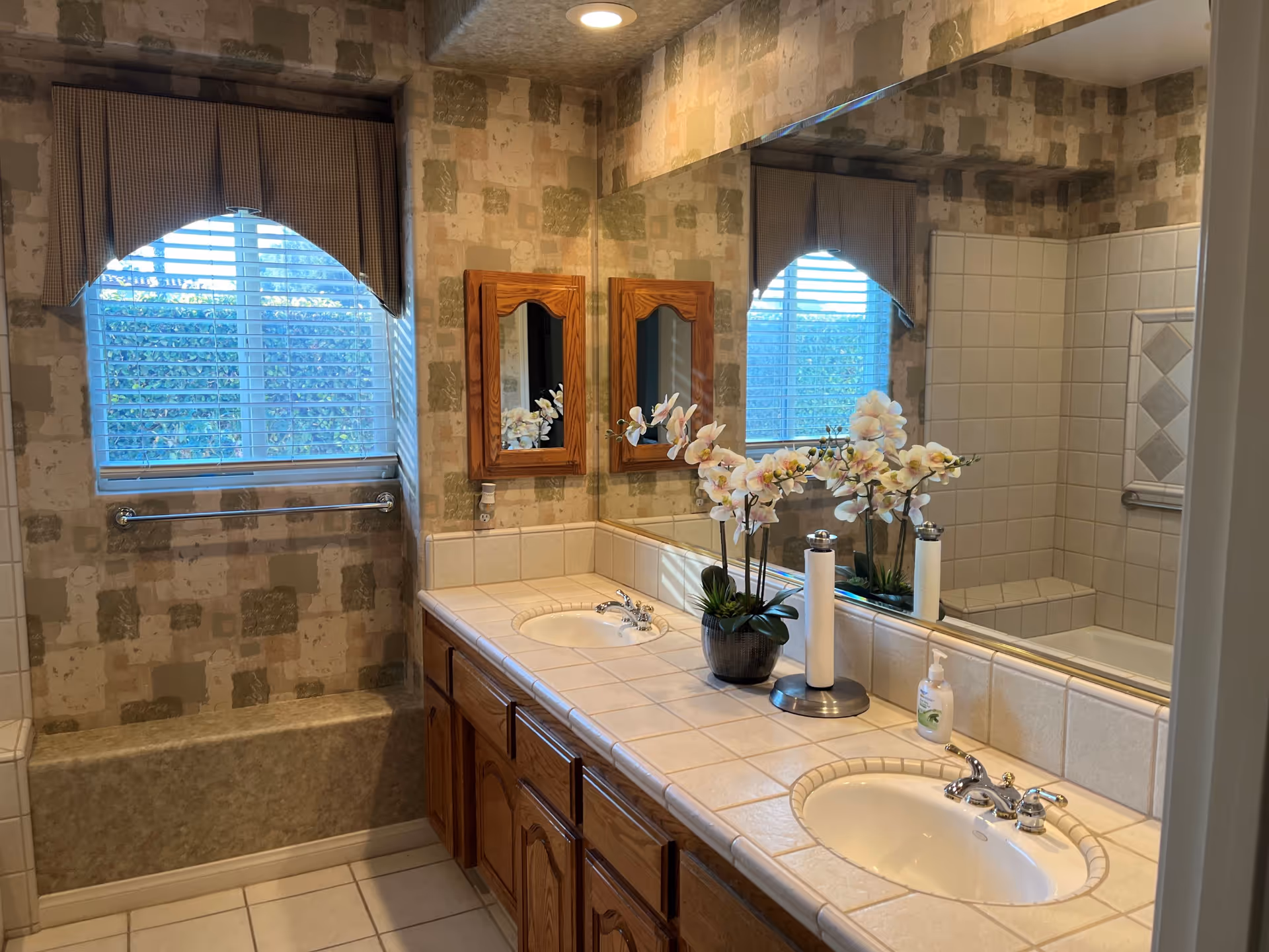 Light-colored bathroom with a double-sink tiled vanity, large mirror, bathtub/shower, and a window with blinds and a valance.