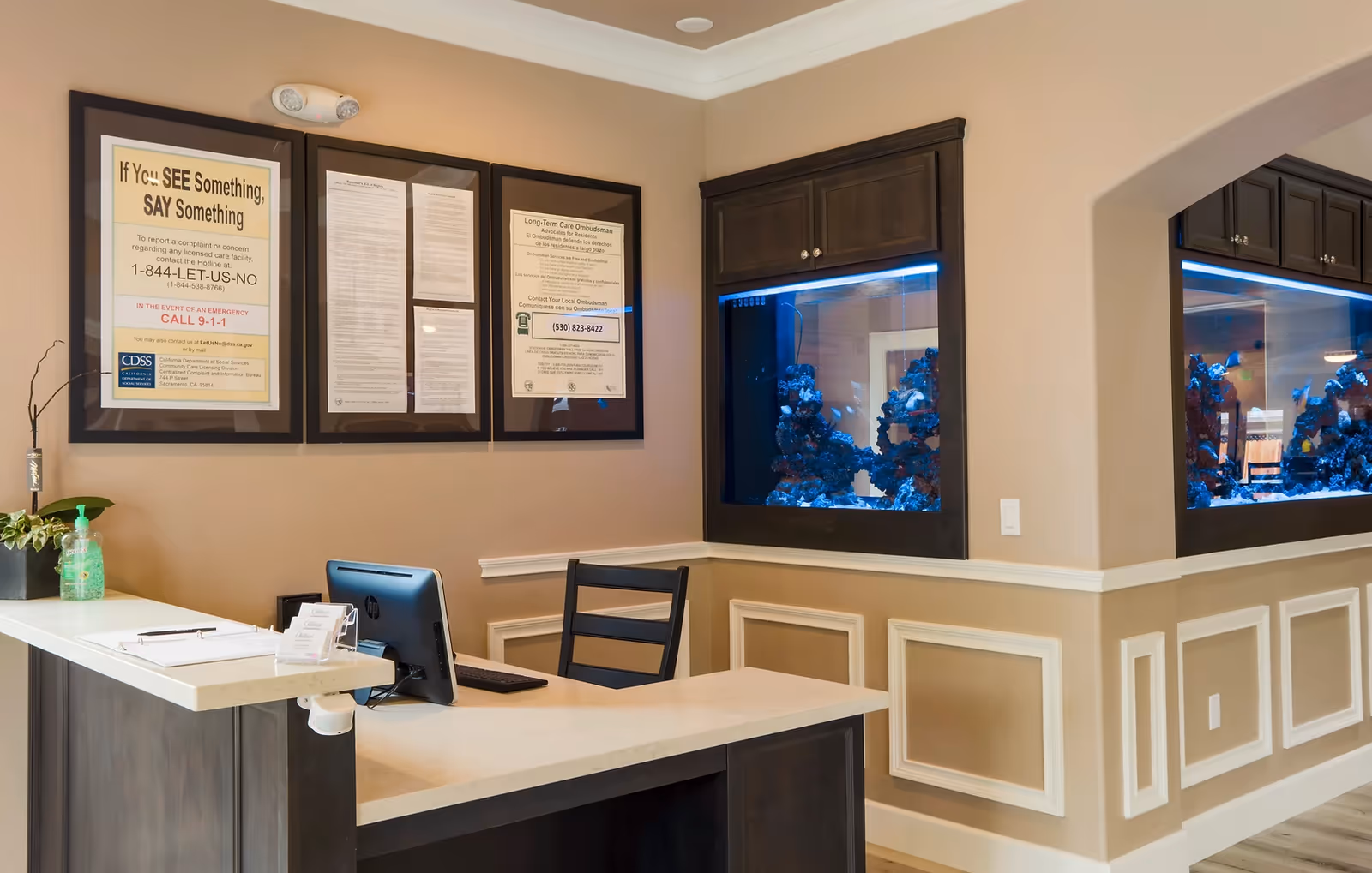 Reception desk area with a computer monitor, chair, and hand sanitizer on the counter. Behind the desk, three framed informational posters are mounted on the wall. To the right, there is a built-in aquarium with blue lighting and decorative coral inside. The walls are beige with white trim and molding details.