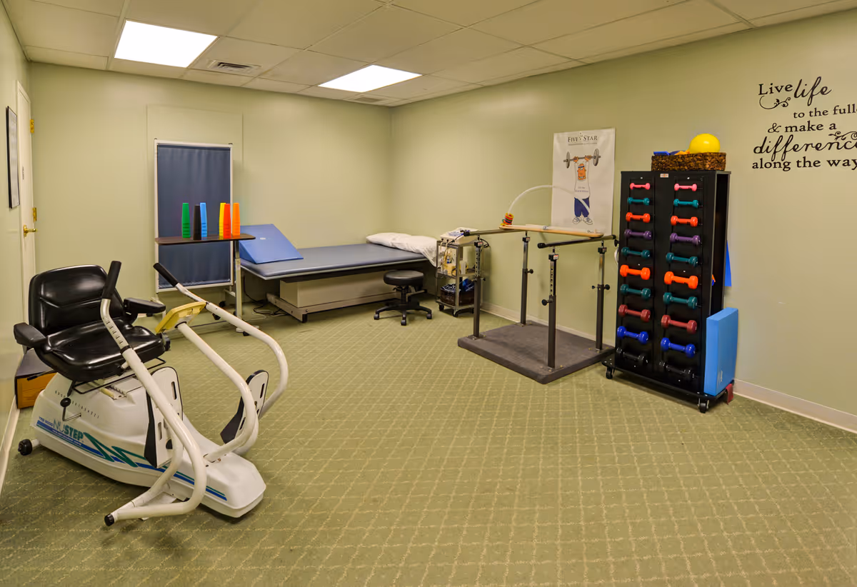 A physical therapy or exercise room with a stationary exercise bike, parallel bars for walking support, a treatment table with a pillow, a rack holding colorful dumbbells, and a small table with stacked plastic cups. The walls are light green, and there is a motivational quote decal on the wall.