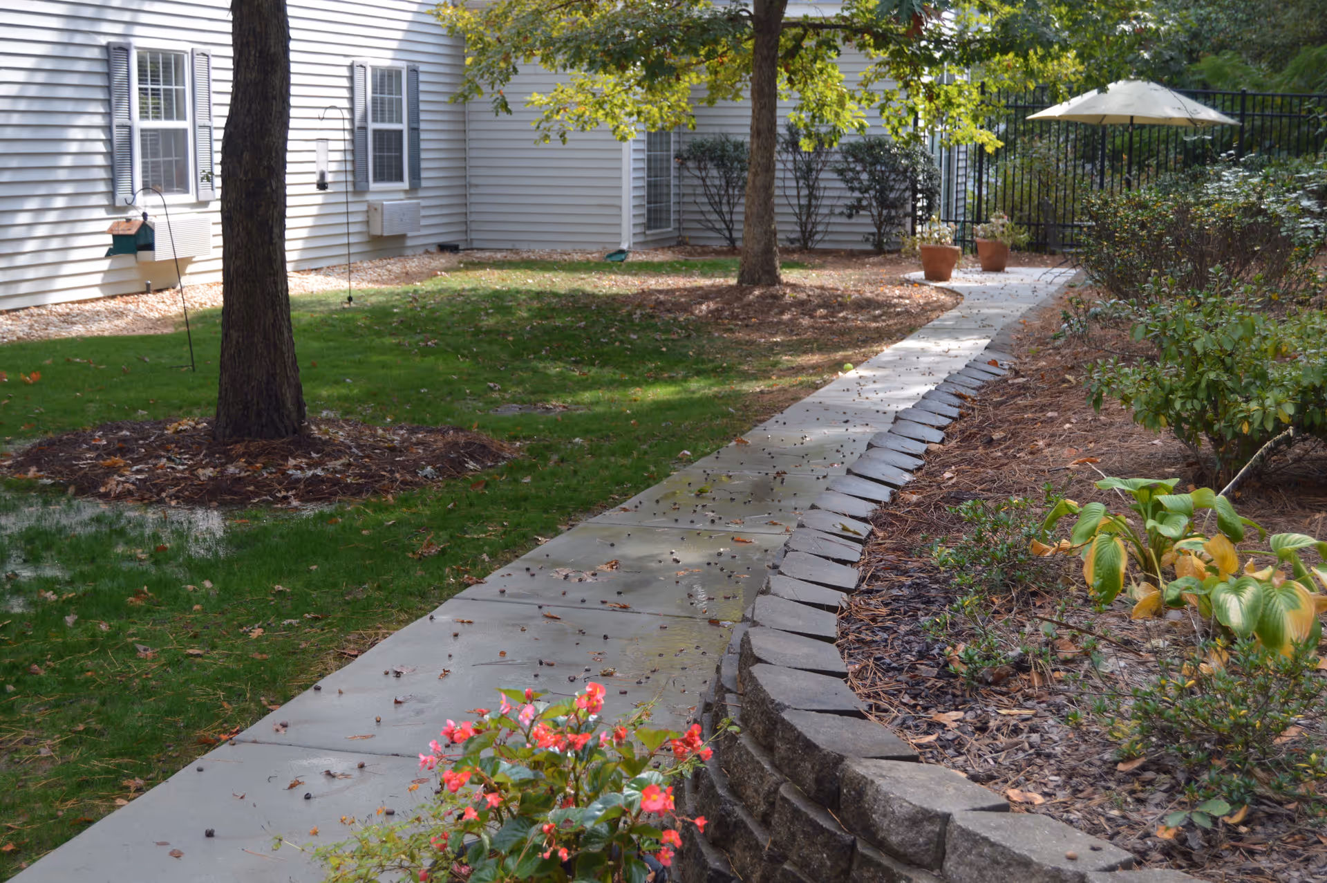 A paved walkway curves through a garden area with green grass, trees, and various plants. There is a building with white siding and multiple windows on the left side. Two potted plants and a white patio umbrella are visible near the end of the walkway.