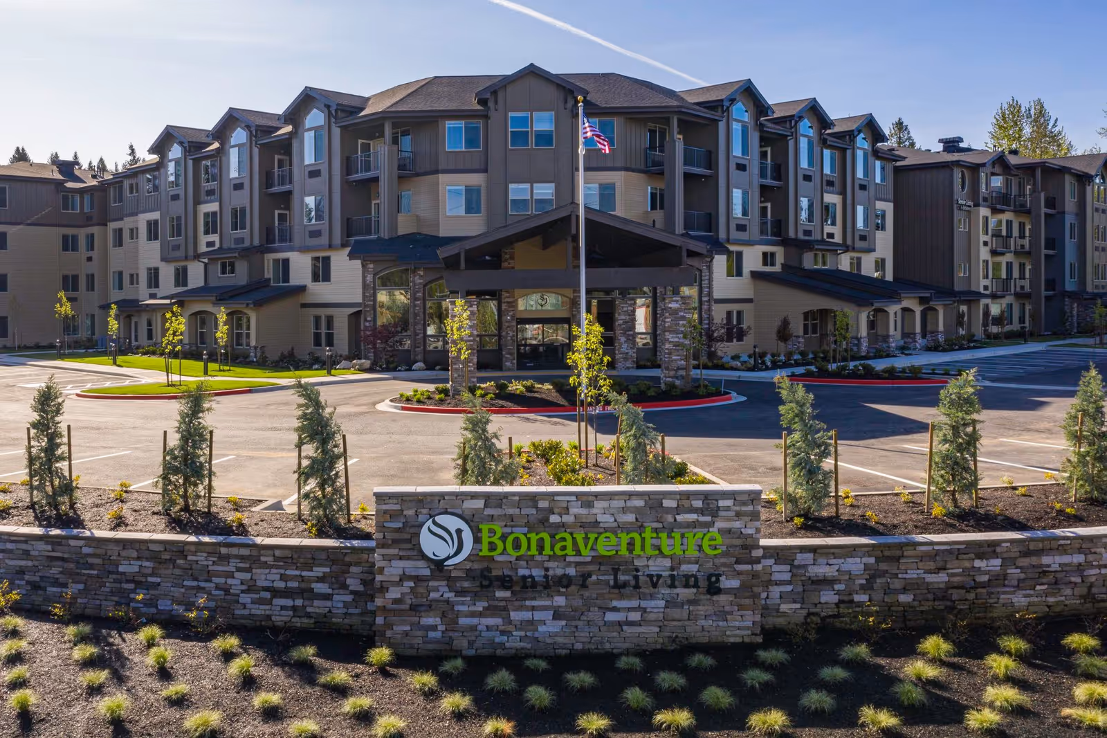 Exterior view of Bonaventure of Puyallup senior living facility showing a large multi-story building with balconies, a covered entrance, a flagpole with an American flag, a parking lot, and landscaped greenery with small trees and shrubs.