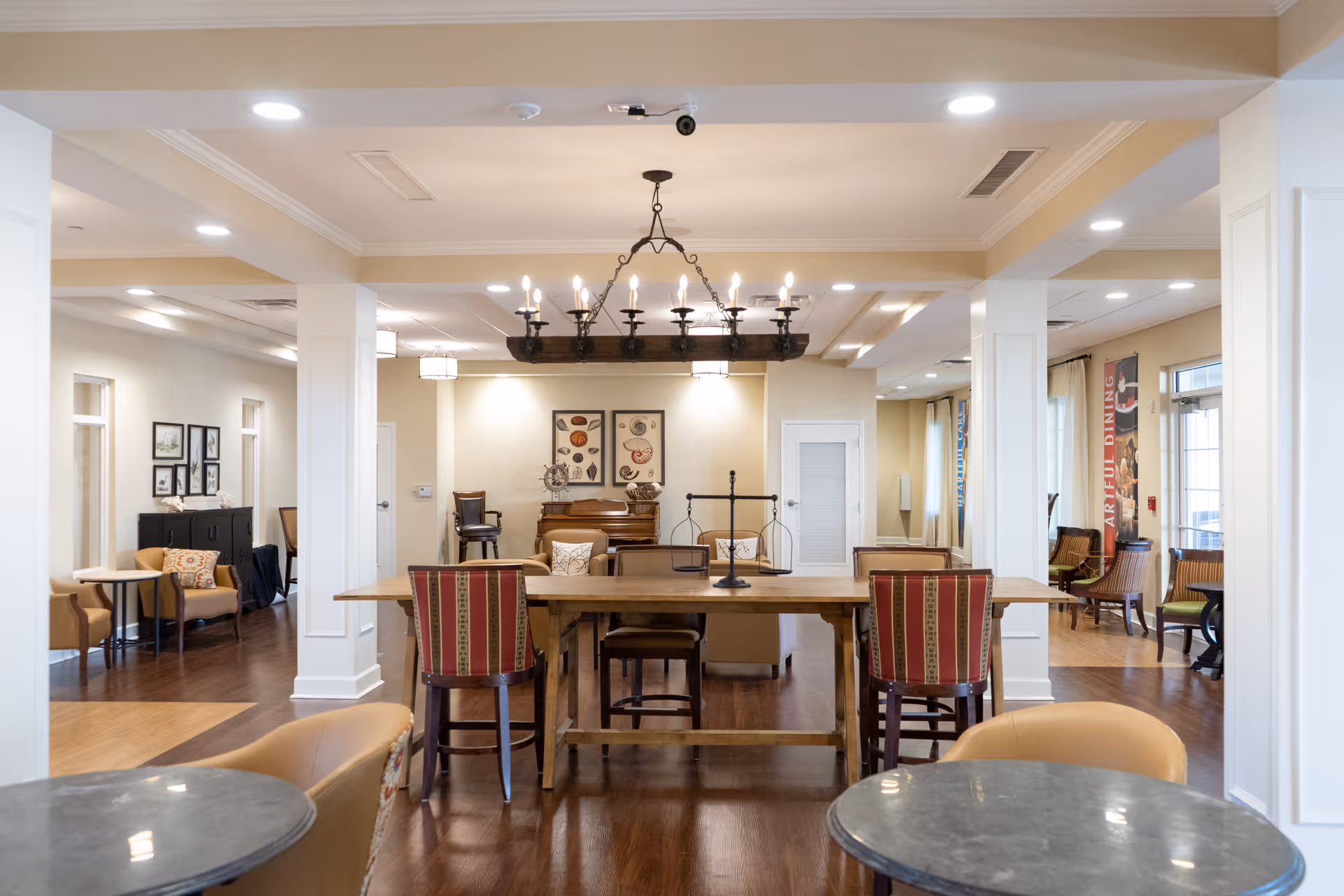 A spacious and well-lit common area in a senior living facility featuring a large wooden table with chairs, comfortable armchairs, and decorative wall art. The room has hardwood floors, white pillars, and a chandelier hanging from the ceiling. Additional seating areas are visible in the background near windows with curtains.