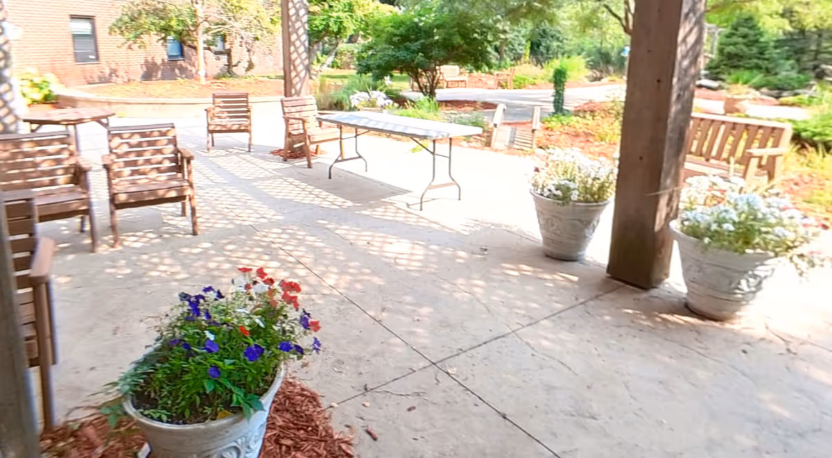 Shaded outdoor patio with wooden chairs, benches, potted flowers, and a folding table under a pergola.