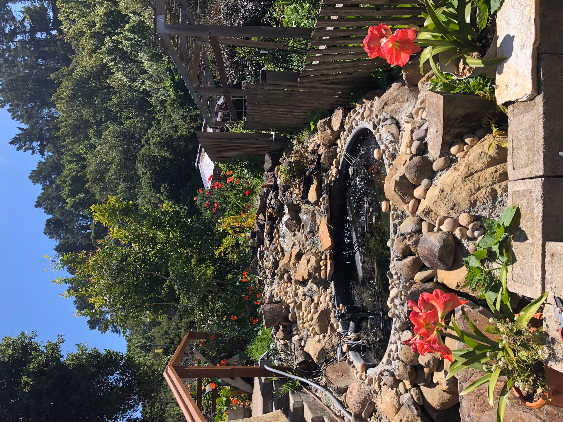 Outdoor garden featuring a stone fountain, rocks, and blooming flowers along a wooden railing.