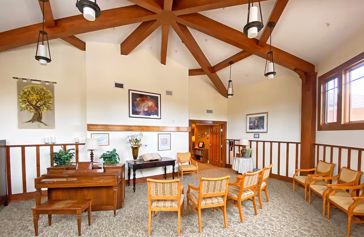 A spacious room with a high ceiling featuring wooden beams and hanging lights. The room contains a wooden piano with a bench, several wooden chairs arranged in a semi-circle, a small table with a vase of flowers, and framed artwork on the walls. Large windows on the right side allow natural light to enter the room.