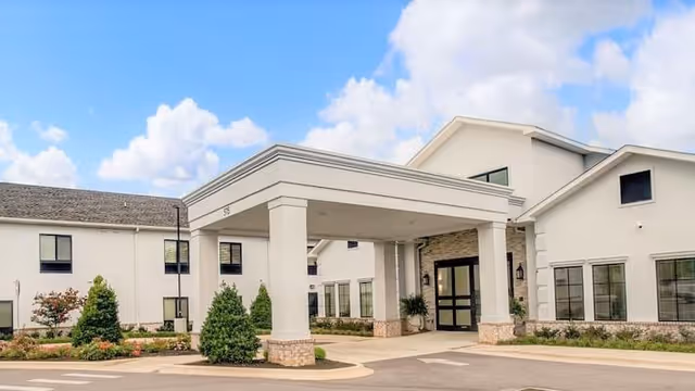 Exterior view of a modern assisted living facility with a covered entrance supported by large white columns, surrounded by landscaped greenery and a clear blue sky with some clouds.
