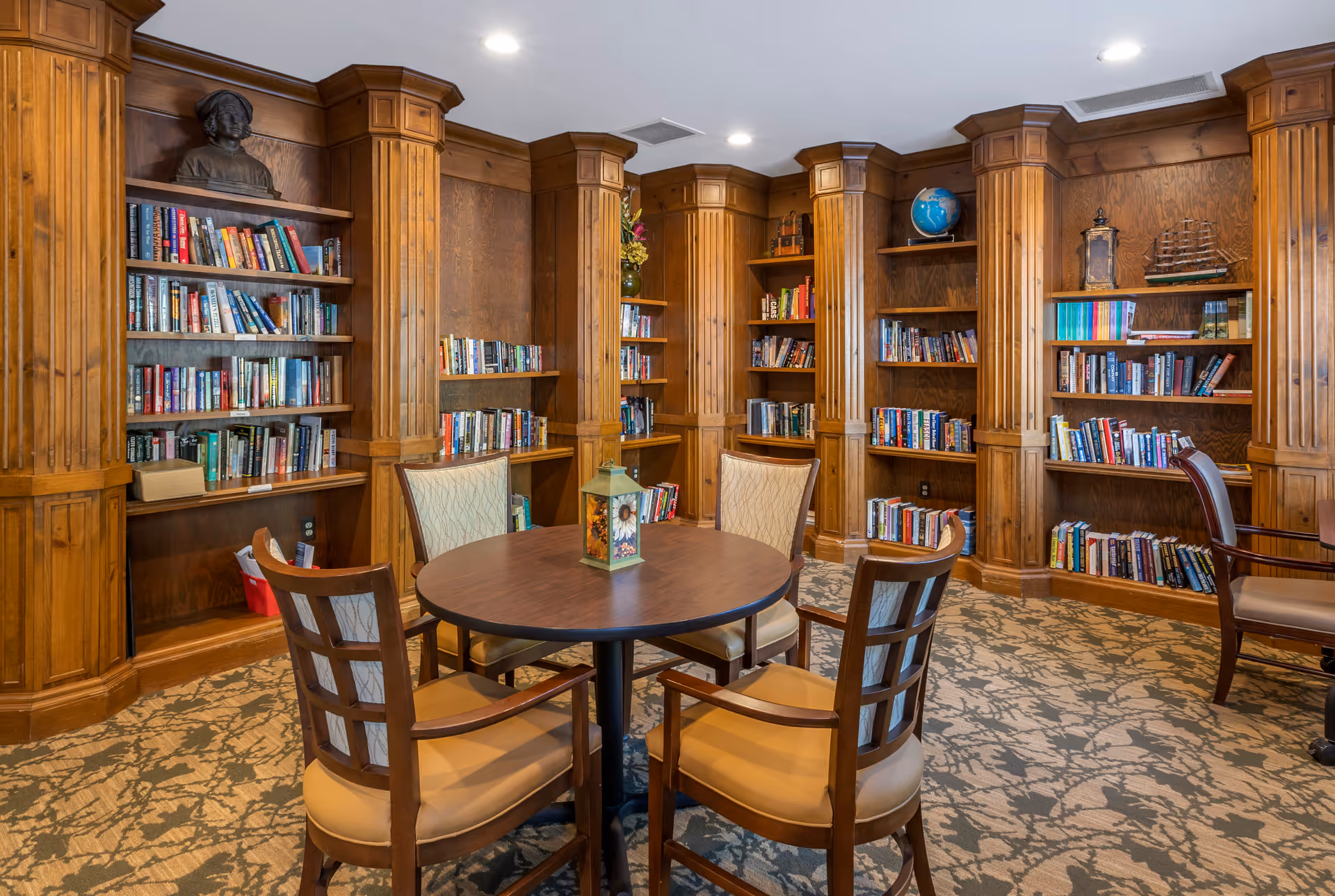 A cozy library room with wooden bookshelves filled with books, a round wooden table in the center surrounded by four cushioned chairs, and decorative items including a bust, a globe, and a model ship on the shelves.