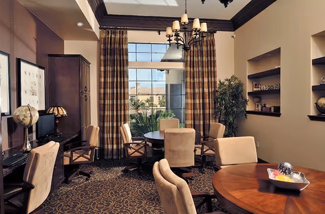 A cozy interior room with multiple round wooden tables surrounded by beige upholstered chairs. There is a large window with plaid curtains letting in natural light, a chandelier hanging from the ceiling, and a computer desk with a globe and lamp on the left side. Shelves with decorative items and a potted plant are on the right wall.